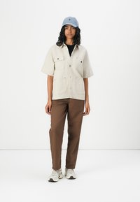 Beige short-sleeve shirt with two chest pockets and black buttons, paired with brown pants and a light blue cap. Sneakers complete the look.