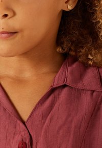Bordeaux shirt met een gestructureerde, gekreukte stof, voorzien van een kraag en een knoopsluiting. Klein gouden studsieraden zichtbaar.