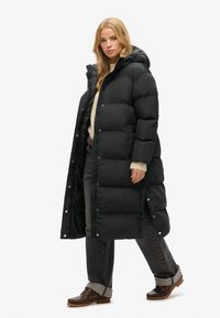 Sort puffet jakke med en lang silhuet, featuring en hætte, trykknapslukninger og sideslids. Bæres over en cremet sweater og grå jeans.