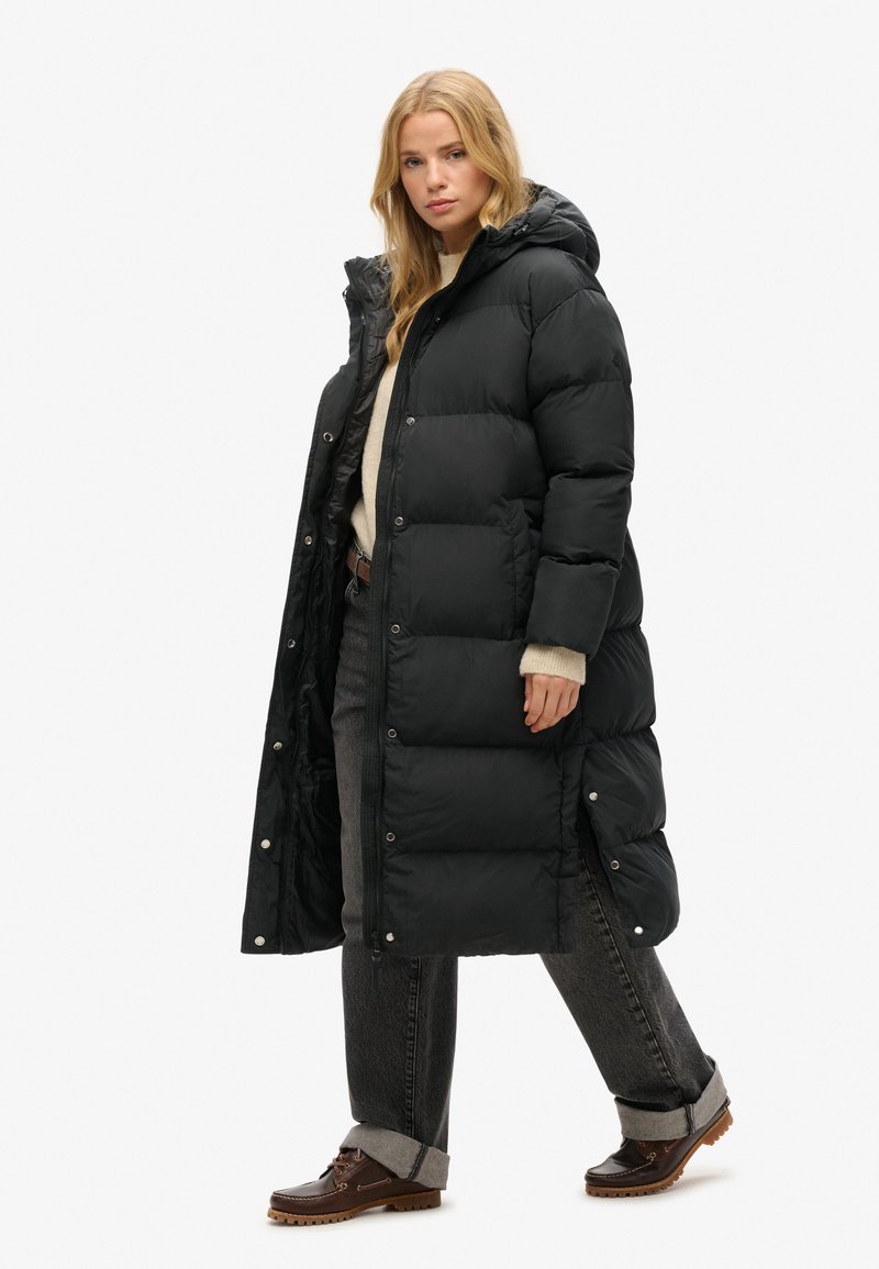 Schwarze Daunenjacke mit langer Silhouette, ausgestattet mit Kapuze, Druckknopfverschlüssen und seitlichen Schlitzen. Getragen über einem cremefarbenen Pullover und grauen Jeans.