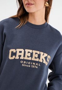 Marineblauwe sweatshirt met ronde hals en geribde mouwuiteinden, met een crèmekleurige "CREEKS" grafiek en de tekst "Original Since 1974".