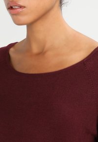 Maglione bordeaux con una scollatura ampia, texture lavorata a maglia e finitura liscia. Il primo piano mette in risalto il materiale e i dettagli della scollatura.