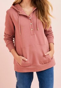 Růžová mikina s kapucí, s částečnou knoflíkovou légou, zlatými doplňky, přední kapsou a žebrovanými manžetami, v kombinaci s džíny.