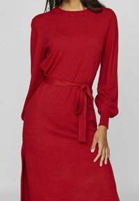 Czerwona dzianinowa sukienka z długimi rękawami, związywanym paskiem w talii i bocznym rozcięciem. Materiał wydaje się miękki, o gładkiej strukturze i dopasowanym kroju.