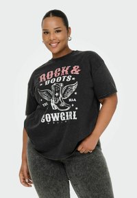 Uśmiechająca się kobieta z spiętymi włosami, nosząca kolczyki w kształcie kół, czarną koszulkę z grafiką "Rock & Boots Cowgirl" oraz wzorem skrzydlatej boot, a także ciemne dżinsy.