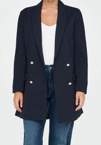 Granatowy podwójnie zapięty blazer z gładkiego materiału, z złotymi guzikami i bocznymi kieszeniami, w zestawieniu z białą bluzką i dżinsami.