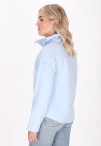 Donna con lunghi capelli biondi, che indossa una giacca azzurro chiaro, si posa di lato su uno sfondo bianco, mettendo in mostra il design della giacca.