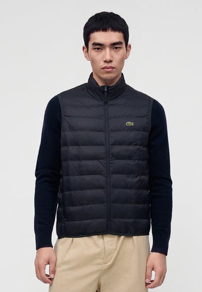 Jeune homme portant une veste matelassée noire zippée sur une chemise à manches longues foncée et un pantalon beige, debout devant un fond gris uni.