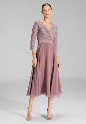 Femme portant une robe mauve à haut en dentelle avec manches trois-quarts et une jupe midi fluide, associée à des talons à bride à la cheville de couleur claire.