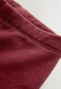 Burgundowy materiał o gładkiej fakturze, z obszytym szwem wzdłuż krawędzi. Materiał wygląda na miękki i lekko elastyczny.