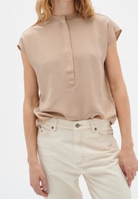 Beige ärmlös blus med mandarin krage, med en frontslå och mjukt, lätt tyg. Kombinerad med ljusa, högmidjade jeans.
