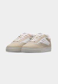 Paire de baskets basses beige et blanches avec lacets et semelles en caoutchouc grises sur un fond clair uni.