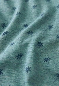 Tissu bleu-gris avec de petits motifs dispersés de palmiers bleu foncé et plis doux visibles ainsi qu'une texture apparente.