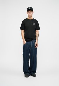 Black t-shirt with a round logo, paired with loose-fitting navy pants featuring white vertical stripes. Complete with a black cap and dark shoes.