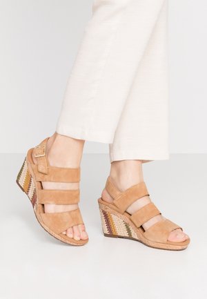 Fødder iført beige ruskinds kilehæle med flerfarvede vævede korkhæle og cremede bukser mod en neutral baggrund.