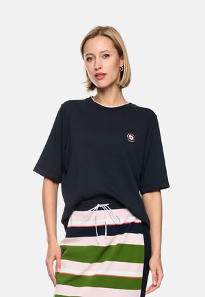 Woman with short blonde hair wearing a loose black t-shirt with flower emblem and a striped skirt with green, pink, and navy bands.