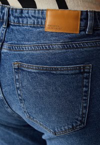 Jeans aus mittlerem Blau mit strukturierter Oberfläche, versehen mit einem braunen Leder-Logo-Patch und klassischen Gesäßtaschen mit sichtbaren Nähten.