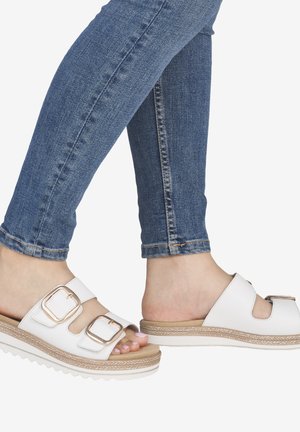 Füße, die weiße Slide-Sandalen mit großen goldenen Schnallen und blaue Skinny-Jeans vor weißem Hintergrund tragen.
