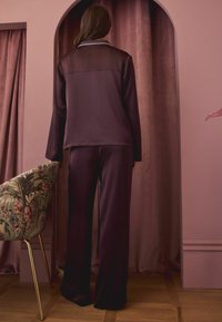 Burgundowa satynowa piżama składająca się z długiego topu z przezroczystym dekoltem oraz dopasowanych spodni z szerokimi nogawkami. W tle widoczny teksturowany kwiatowy fotel.