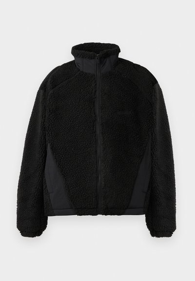 Casaco de polar preto com gola alta, apresentando um exterior texturizado e peludo, e detalhes de tecido suave nas laterais. Fecho frontal com zíper.