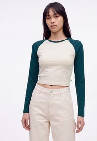 Jeune femme aux cheveux raides et foncés portant un haut court à manches longues crème et vert et un pantalon beige taille haute, debout.