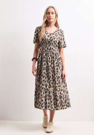Femme aux longs cheveux blonds portant une robe midi beige et noire à motif léopard et des baskets blanches, debout devant un fond clair uni.