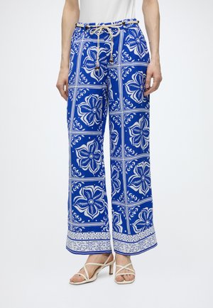 Pantalones anchos azules con patrones de azulejos florales blancos y un cinturón de cuerda, combinados con sandalias blancas de tacón con tiras.
