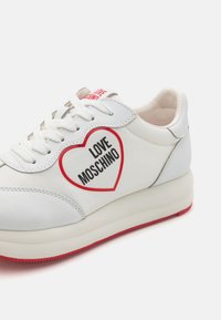 Fehér sneaker piros részletekkel és szív alakú logóval. Simán bőr és textil anyagokból készült, lapos fűzőkkel és párnázott talppal.