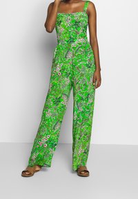 Jumpsuit floreale verde con gambe ampie, scollatura quadrata e spalline sottili. Presenta vivaci motivi di foglie e fiori. Indossati sandali.