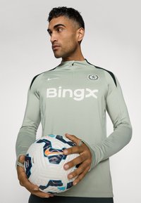 Langärmliges, hellgrünes Nike Trainingstop mit dunkelgrünen Akzenten und bedrucktem Logo, kombiniert mit einem gemusterten weißen und blauen Fußball.