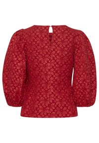 Blouse rouge texturée avec un motif floral, manches bouffantes et une fermeture à l'arrière en forme de trou de serrure. Surface lisse avec une coupe structurée.