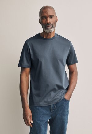 Erwachsener Mann mit grauem Bart, der ein schlichtes dunkelgraues T-Shirt und blaue Jeans trägt, steht mit einer Hand in der Tasche vor neutralem Hintergrund.