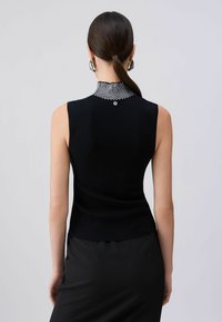 Femme aux cheveux foncés attachés en queue de cheval, portant des boucles d'oreilles argentées en forme de créoles et un haut noir sans manches avec un col haut à motifs, vue de dos.