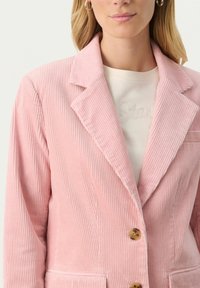 Rosa kordflorsblazer med en notch lapel, två framfickor och knappstängning. Har vertikal ribbad textur och ljusbruna knappar.