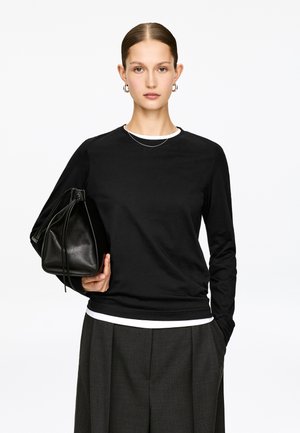 Jonge vrouw met naar achteren gestyled haar, gekleed in een zwart shirt met lange mouwen, grijze broek, zilveren creolen en een zwarte leren handtas vasthoudend.