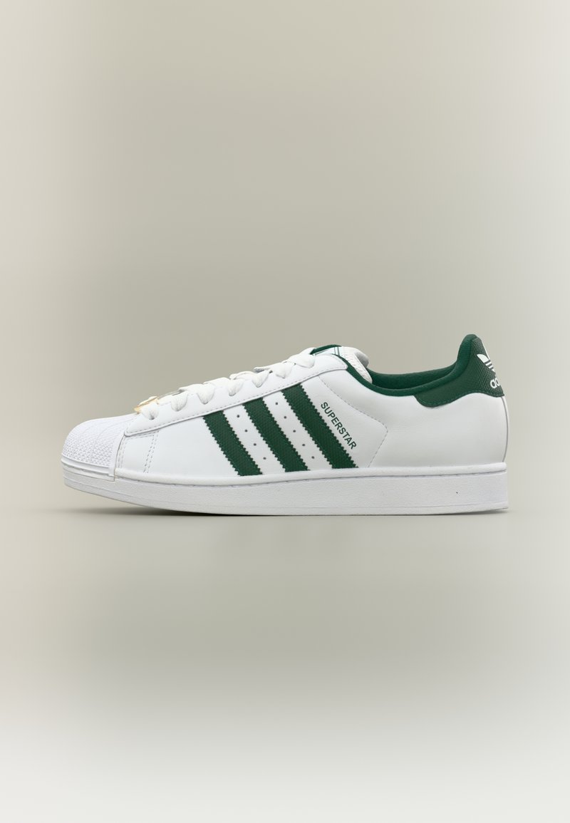 Witte lederen sneaker met groene accenten, voorzien van drie strepen aan de zijkant, een rubberen neuskap en platte veters. De tekst luidt "SUPERSTAR."