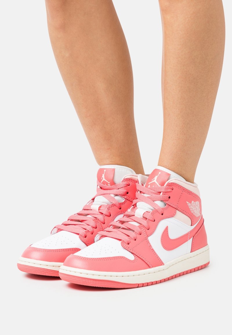 Pieds portant des baskets montantes Nike Air Jordan roses et blanches avec les lacets noués, présentés sur un fond clair uni.
