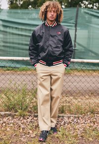 Black bomber jacket with red and white striped cuffs, beige pants, and black sneakers with pink accents, standing in front of a fence.