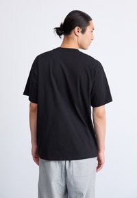 Mand med samlet hår iført en simpel, sort t-shirt og lysegrå bukser, vendt væk fra kameraet mod en hvid baggrund.