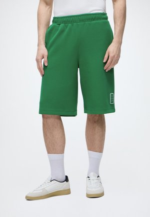 Homme portant un short vert au genou, des chaussettes blanches, des baskets blanches et une chemise blanche sur un fond uni.