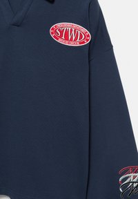 Pullover maniche lunghe blu navy con scollo a V, logo ricamato in rosso e bianco e dettagli decorativi con fili multicolori sulla manica.