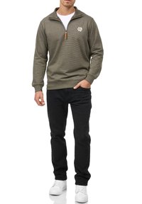 Olivengrøn zip-up sweatshirt med et hvidt logo, ribbede ærmer og krave. Kombineret med sorte bukser og hvide sneakers. Glat tekstur.