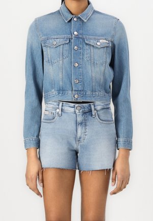 Osoba ubrana w jasno-niebieską, krótką kurtkę jeansową zapinaną na guziki oraz pasujące, wysokowęziowe szorty jeansowe z postrzępionymi brzegami.