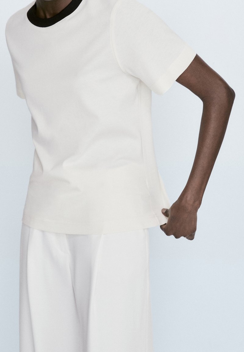 Personne portant une chemise blanche à manches courtes avec un col noir et un pantalon blanc, ajustant l'ourlet latéral de la chemise.