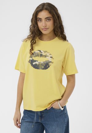 Kvinde med krøllet hår iført en gul T-shirt med et gråt og sort camouflage læbeprint, blå jeans og et sølvarmbånd, hånd i lommen.