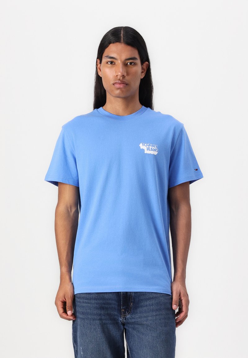 T-shirt en coton bleu clair à manches courtes et col rond. Arbore un logo blanc sur la poitrine gauche ; porté avec un jean noir.