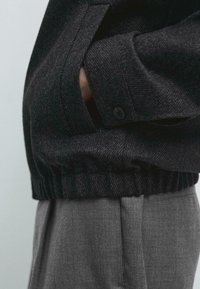 Czarna, fakturowana kurtka z prążkowanym dołem i boczną kieszenią, zestawiona z jasnoszarymi, dopasowanymi spodniami o płaskim przodzie i subtelnym kantem.