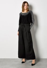 Donna che indossa pantaloni neri a gamba larga, una maglietta nera a maniche lunghe con dettagli argentati sul colletto e sulle maniche, e scarpe con tacco alto nere.