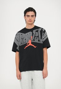 Μαύρο βαμβακερό T-shirt με μεγάλο λευκό κείμενο “Jordan” και κόκκινο περίγραμμα ενός άλτη. Κοντά μανίκια και λαιμόκοψη τύπου crew.