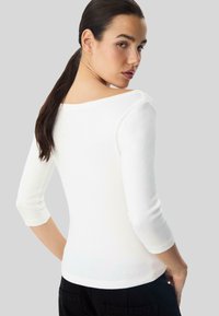 Weißes enges Oberteil mit Dreiviertelärmeln, weichem Material und weitem Ausschnitt. Das Modell zeigt den Rücken und hebt das Rückendesign hervor.